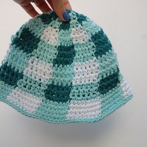 Op de afbeelding: Een handgemaakte gehaakte emmerhoed in een dambordpatroon. De hoed heeft vierkanten van wit, teal en donker teal. De rand en de rand zijn effen teal van kleur. De hoed wordt vastgehouden door een hand met blauwe nagellak.