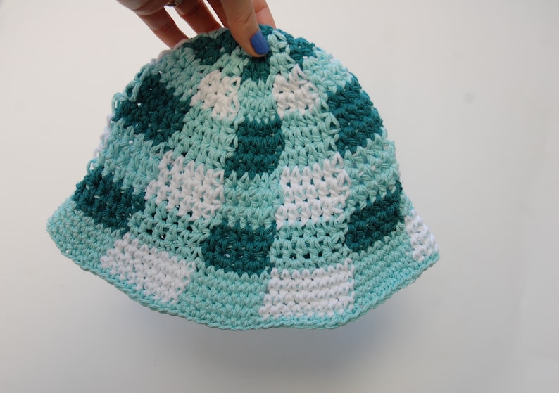 Op de afbeelding: Een handgemaakte gehaakte emmerhoed in een dambordpatroon. De hoed heeft vierkanten van wit, teal en donker teal. De rand en de rand zijn effen teal van kleur. De hoed wordt vastgehouden door een hand met blauwe nagellak.