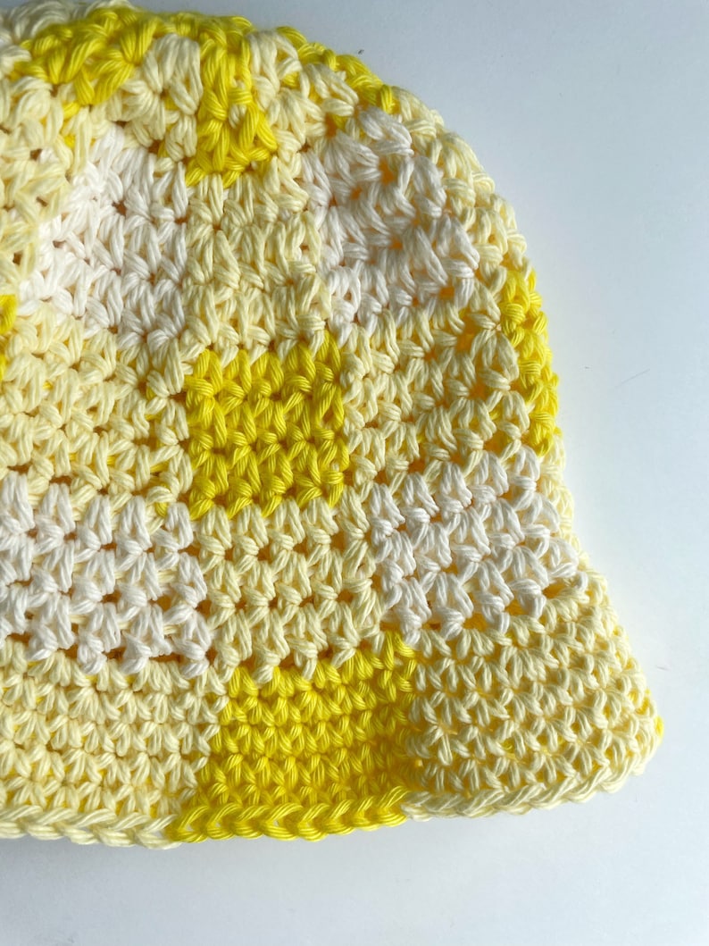 Op de afbeelding: Een handgemaakte gehaakte bucket hat in een levendig geel en cr&egrave;me kleurenschema. De hoed heeft een textuur steekpatroon en een klassieke bucket hat vorm. De hoed is gemaakt van zacht garen, perfect voor zonnige dagen.