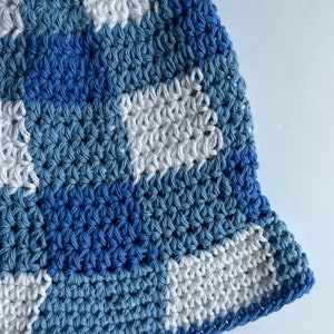 Op de afbeelding: Een handgemaakte gehaakte vissershoed in tinten blauw en wit. De hoed heeft een geruit patroon met verschillende tinten blauw en wit, waardoor een textuur ontstaat. De rand is effen donkerblauw. Deze hoed is een stijlvol accessoire.