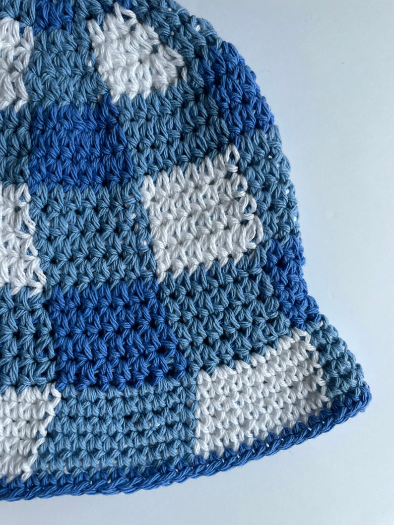 Op de afbeelding: Een handgemaakte gehaakte vissershoed in tinten blauw en wit. De hoed heeft een geruit patroon met verschillende tinten blauw en wit, waardoor een textuur ontstaat. De rand is effen donkerblauw. Deze hoed is een stijlvol accessoire.