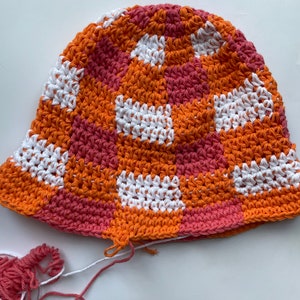Op de afbeelding: Een handgemaakte gehaakte bucket hat met een patchwork design. De hoed heeft vierkanten van oranje, witte en roze garen. Los garen in roze en wit is zichtbaar bij de hoed. Deze hoed is een stijlvol accessoire.