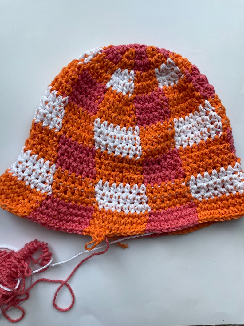 Op de afbeelding: Een handgemaakte gehaakte bucket hat met een patchwork design. De hoed heeft vierkanten van oranje, witte en roze garen. Los garen in roze en wit is zichtbaar bij de hoed. Deze hoed is een stijlvol accessoire.