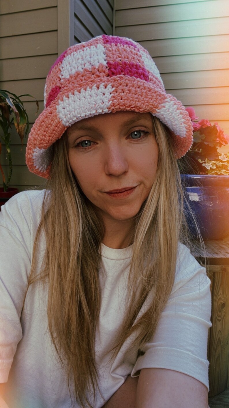 Op de afbeelding: Een handgemaakte gehaakte bucket hat in een dambordpatroon van wit, roze en perzik. De hoed heeft een brede rand en wordt gedragen door een persoon met lang blond haar en een wit t-shirt.