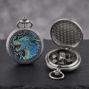 Peut inclure: Une montre de poche argentée avec un motif de dragon et un compartiment contenant des dés. Le dragon est représenté dans des tons de bleu et de vert sur un fond d'éclairs. La montre est ouverte, révélant les dés.