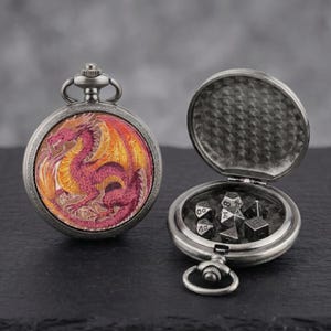 May include: A silver pocket watch featuring a dragon design on the face. The watch is open, revealing dice inside. The dragon is red and orange, set against a yellow background. The watch is on a black surface.