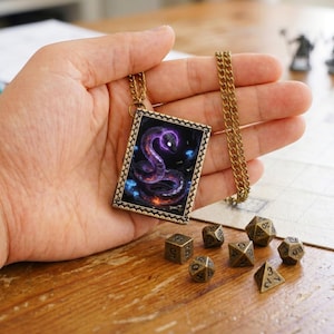 Può includere: Una collana con ciondolo rettangolare raffigurante un'illustrazione scura di un serpente viola brillante avvolto tra funghi blu, incastonata in una cornice color bronzo con catenina abbinata, accompagnata da sette dadi poliedrici color bronzo.