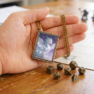 Puede incluir: Un collar con colgante rectangular que muestra una ilustración de un dragón blanco y verde azulado sobre fondo morado, enmarcado en bronce texturizado con cadena de eslabones a juego y siete dados de juego poliédricos de bronce.