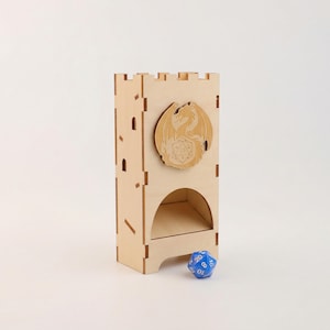 Puede incluir: Una torre de dados de madera clara con un emblema de dragón y un dado azul de veinte caras. La torre, con diseño de castillo, presenta ventanas recortadas y una abertura curva para lanzar los dados. El emblema del dragón es un relieve.