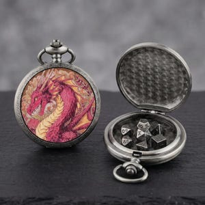 Puede incluir: Un reloj de bolsillo plateado con un dragón rojo en la parte delantera. El reloj está abierto, revelando un conjunto de dados. El reloj está sobre una superficie oscura.