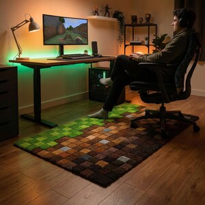 May include: A pixelated rug in shades of green, brown, and gray, resembling a video game landscape, sits on a wooden floor. The rug is placed in front of a desk with a computer setup, suggesting a gaming or work area.