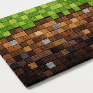 May include: A rectangular doormat with a pixelated design. The top section is green, transitioning to brown and dark brown squares. Several gray squares are interspersed throughout the design. The mat is made of a textured material.