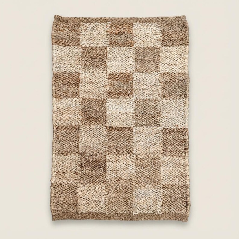 May include: A rectangular, woven rug with a checkerboard pattern. The squares alternate between light beige and various shades of brown. The rug has a textured surface and a natural, earthy aesthetic.