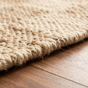 May include: Close-up of a natural fiber rug with a woven texture. The rug is a light tan color and appears to be made of jute or a similar material. The edges of the rug are visible, showing the thickness and construction.
