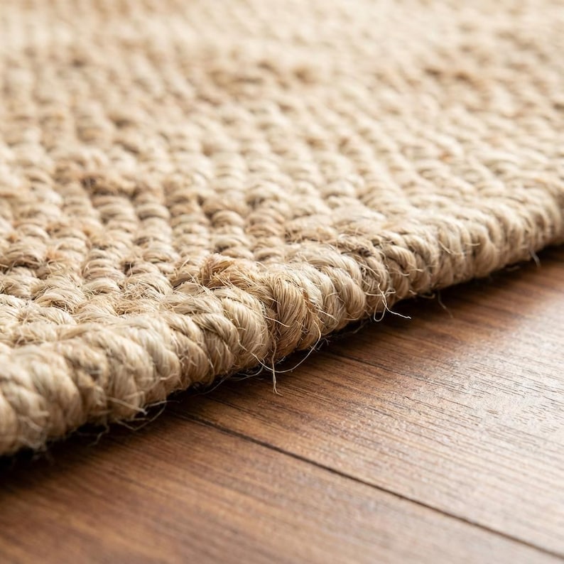 May include: Close-up of a natural fiber rug with a woven texture. The rug is a light tan color and appears to be made of jute or a similar material. The edges of the rug are visible, showing the thickness and construction.