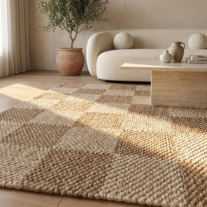 May include: A woven area rug with a checkerboard pattern in shades of beige and tan. The rug is made of natural fibers and has a textured appearance. The rug is placed in a living room setting with a cream-colored sofa and a wooden coffee table.