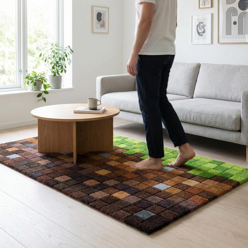 May include: A rectangular area rug with a pixelated design in shades of brown and green. The rug is placed in a living room setting with a wooden coffee table and a gray sofa. The rug's design resembles a block pattern.