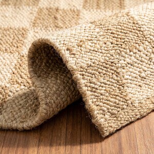 May include: Close-up of a rolled-up, natural-toned jute rug. The rug has a woven texture with a subtle checkered pattern. The edges are neatly finished, and the rug sits on a wooden surface.