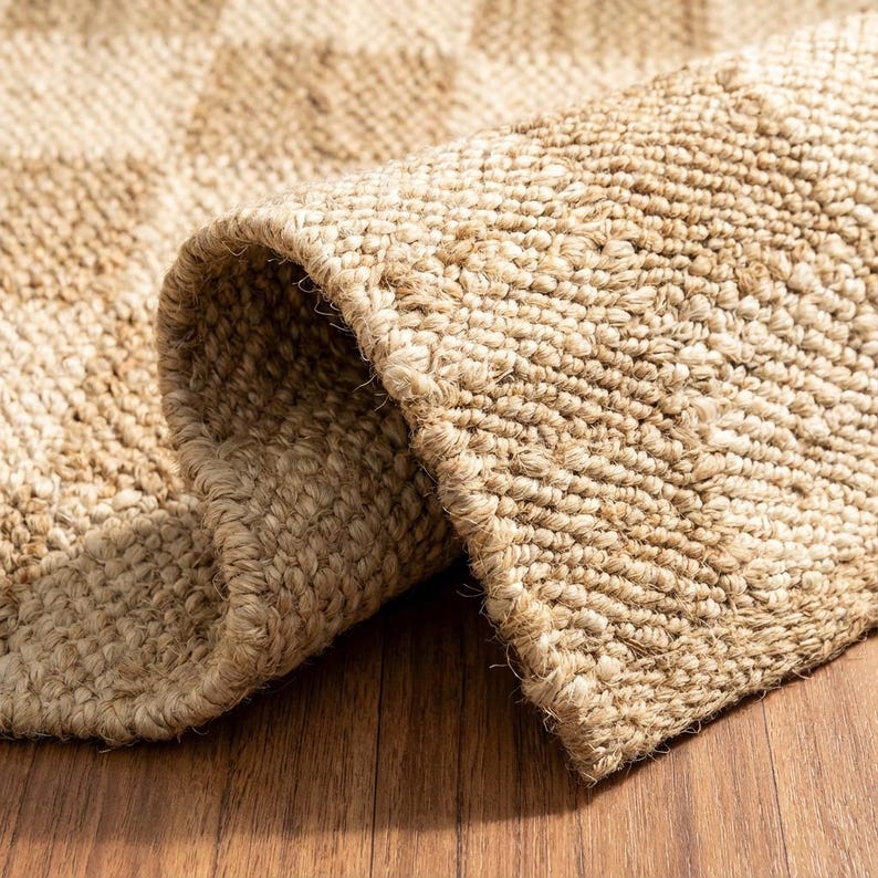 May include: Close-up of a rolled-up, natural-toned jute rug. The rug has a woven texture with a subtle checkered pattern. The edges are neatly finished, and the rug sits on a wooden surface.
