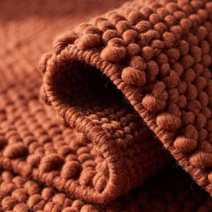 Op de afbeelding: Een terracotta-kleurig kleed met een dik, getextureerd weefsel met verhoogde, afgeronde lussen. De stof is omgevouwen om de dichte, geknoopte constructie van het materiaal te onthullen, met een consistente diepe oranjebruine tint.