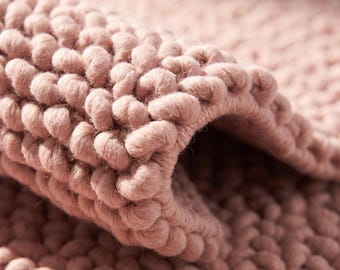May include: A dusty rose colored rug featuring a thick, chunky knit texture with large, interconnected yarn loops. The material is folded over to reveal the dense, consistent weave pattern and the soft, rounded appearance of the individual stitches.