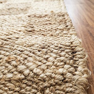 May include: Close-up of a natural fiber rug with a woven texture. The rug is a light tan color and appears to be made of jute or a similar material. The edge of the rug is visible, along with a section of a wooden floor.