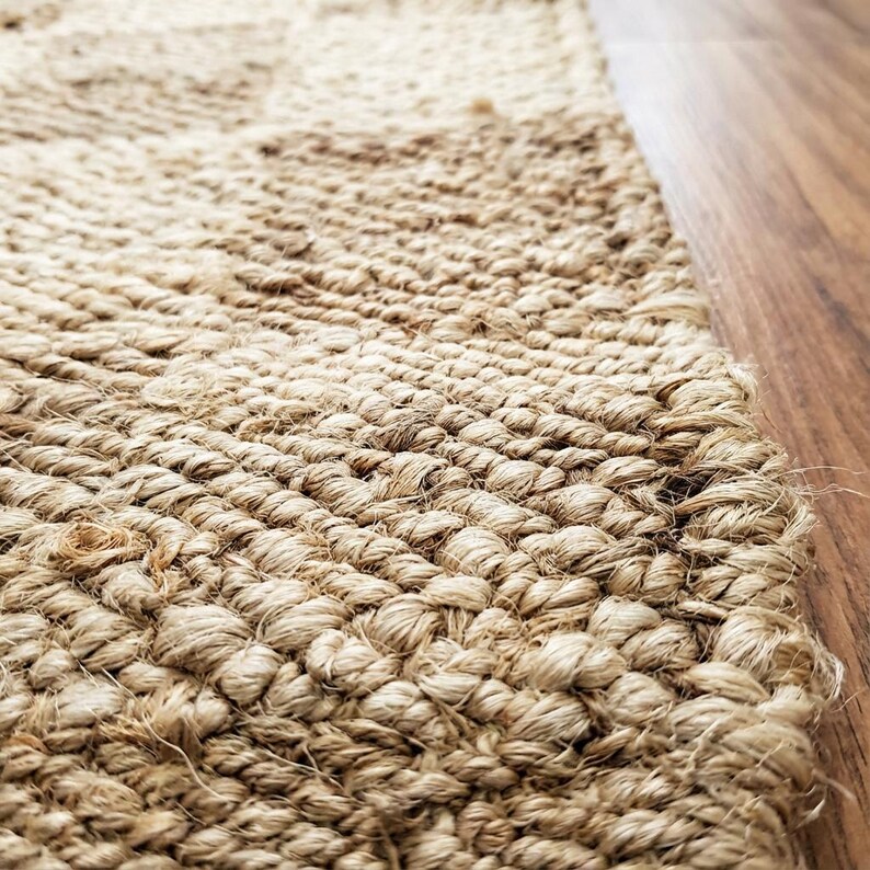 May include: Close-up of a natural fiber rug with a woven texture. The rug is a light tan color and appears to be made of jute or a similar material. The edge of the rug is visible, along with a section of a wooden floor.