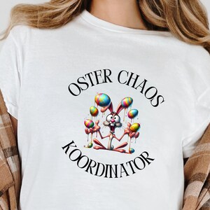 Könnte beinhalten: Weißes T-Shirt mit dem Text "OSTER CHAOS KOORDINATOR" und einer Cartoon-Hasen-Grafik, die bunte Luftballons hält. Der Hase trägt eine Krone und hat einen schelmischen Ausdruck. Ein lässiges Kleidungsstück für den Alltag.