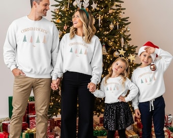 Puede incluir: Una familia con sudaderas blancas a juego con el texto "CHRISTMASCREW" y gráficos de árboles de Navidad coloridos. La familia está de pie frente a un árbol de Navidad decorado, tomados de la mano. Las sudaderas tienen un tema festivo.