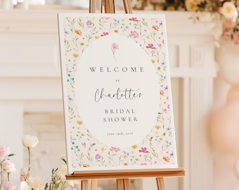 May include: A floral-themed welcome sign on a wooden easel. The sign reads "Welcome to Charlotte's Bridal Shower" with the date June 25th, 2024. The sign is surrounded by colorful flowers and is set in a bright room.