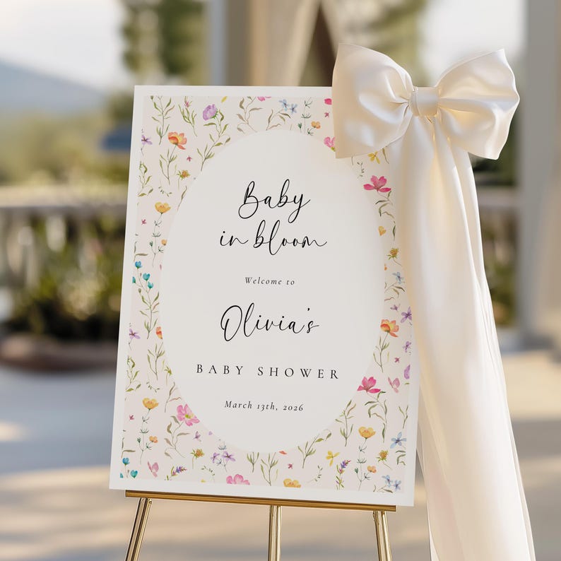 May include: A baby shower welcome sign with the text "Baby in bloom" and "Welcome to Olivia's". The sign is decorated with a floral border and a large cream-colored bow. The sign is on a gold easel.