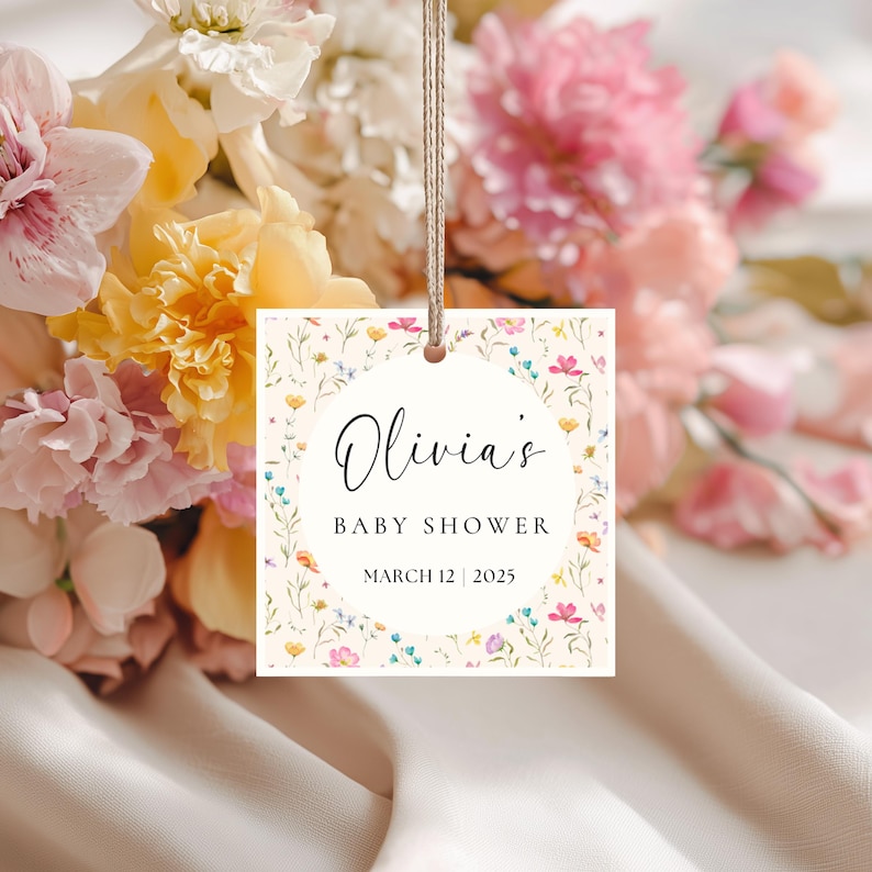 May include: A square baby shower tag with floral accents and the text "Olivia's BABY SHOWER MARCH 12 | 2025". The tag is cream-colored with a floral border and is hanging from a string. A bouquet of flowers is in the background.