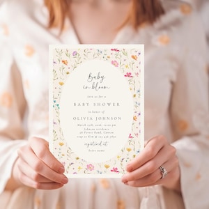 Może przedstawiać: Zaproszenie na baby shower z kwiatowym wzorem. Zaproszenie zawiera tekst "Baby in bloom" oraz szczeg&oacute;ły wydarzenia, w tym datę, godzinę i miejsce. Zaproszenie jest trzymane przez osobę.