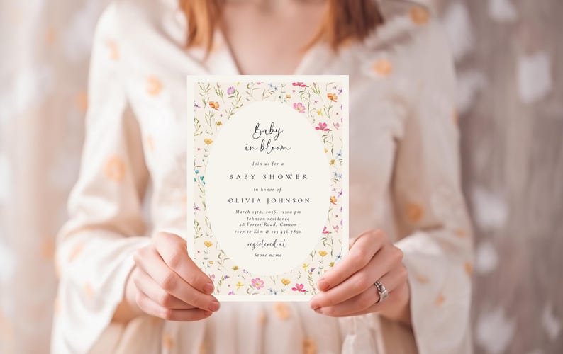 Może przedstawiać: Zaproszenie na baby shower z kwiatowym wzorem. Zaproszenie zawiera tekst "Baby in bloom" oraz szczeg&oacute;ły wydarzenia, w tym datę, godzinę i miejsce. Zaproszenie jest trzymane przez osobę.