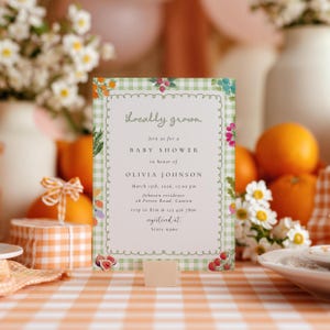 May include: A baby shower invitation with a green gingham border and floral illustrations. The text reads "Locally grown" and details for Olivia Johnson's baby shower, including the date, time, and location. The invitation is propped up on a table.