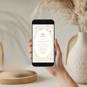 May include: A smartphone displays a baby shower invitation. The invitation features an oval frame with colorful floral accents. The text reads "Baby in bloom" and includes details for Olivia Johnson's baby shower.