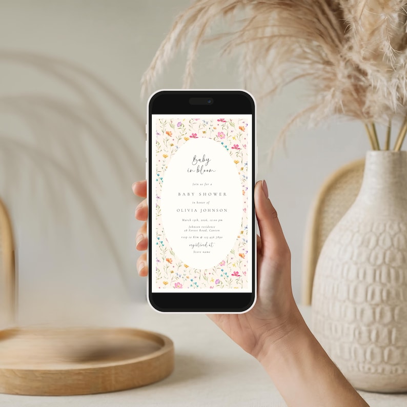May include: A smartphone displays a baby shower invitation. The invitation features an oval frame with colorful floral accents. The text reads "Baby in bloom" and includes details for Olivia Johnson's baby shower.