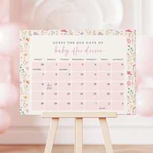 May include: A pink and floral baby shower calendar on a wooden easel. The calendar reads "Guess the Due Date of baby Henderson" and includes dates from November to January. The calendar is decorated with floral accents.