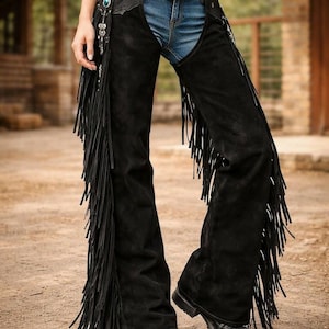 May include: Black suede chaps with long fringe detailing. The chaps are worn over blue jeans and a decorative belt with a large buckle. The outfit is completed with black boots, creating a Western-inspired look.