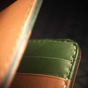 May include: Close-up of a handcrafted leather wallet. The wallet features a tan exterior and a green interior with visible card slots. The edges are meticulously stitched with light green thread, showcasing the craftsmanship. The background is dark.