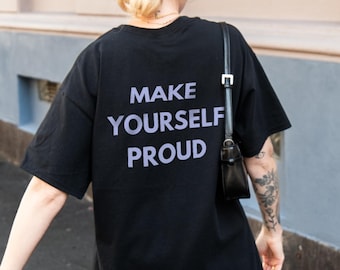 May include: Black oversized t-shirt with the text "MAKE YOURSELF PROUD" in white. The person is wearing a white lace skirt, grey trainers, and a brown baseball cap. A black handbag hangs from the shoulder.