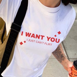 Może przedstawiać: Biała koszulka z czerwonym napisem "I WANT YOU I JUST CAN'T FLIRT". Osoba ma na sobie jasnoniebieskie dżinsy i szare trampki. Widoczny jest brązowy plecak z czarnym paskiem. W dłoni trzymany jest kubek z kawą.