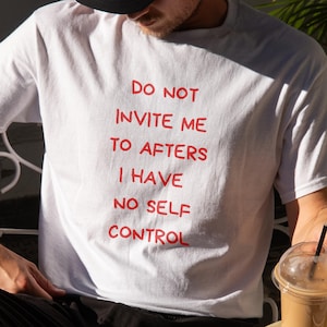 May include: White t-shirt with the red text "DO NOT INVITE ME TO AFTERS I HAVE NO SELF CONTROL." The person is wearing a black cap and sitting at a table with a drink and a croissant. An open book is on the table.