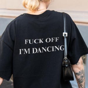 Könnte beinhalten: Schwarzes kurzärmeliges T-Shirt mit dem weißen Text "FUCK OFF I'M DANCING". Die Person trägt eine braune Kappe und einen weißen Spitzenrock. Eine schwarze Handtasche hängt über der Schulter.