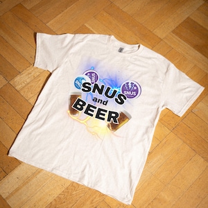 Puede incluir: Camiseta de color crema con un diseño gráfico. El diseño presenta las palabras "SNUS and BEER" en negrita, dos vasos de cerveza y logotipos circulares morados con la palabra "SNUS".