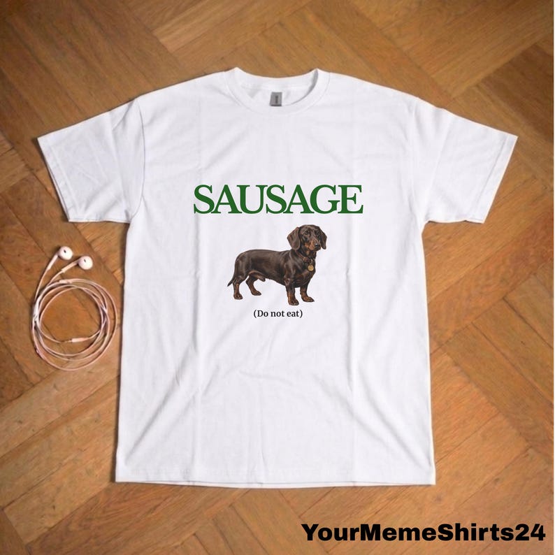 May include: White t-shirt with the word "SAUSAGE" in green above a brown dachshund dog illustration. Below the dog is the text "(Do not eat)". The shirt is on a wooden surface.