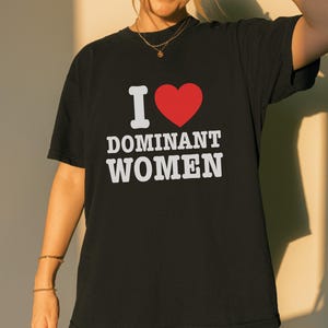 Puede incluir: Camiseta negra con el texto "I ♥ DOMINANT WOMEN" en blanco y un símbolo de corazón rojo. La camiseta es de estilo casual.