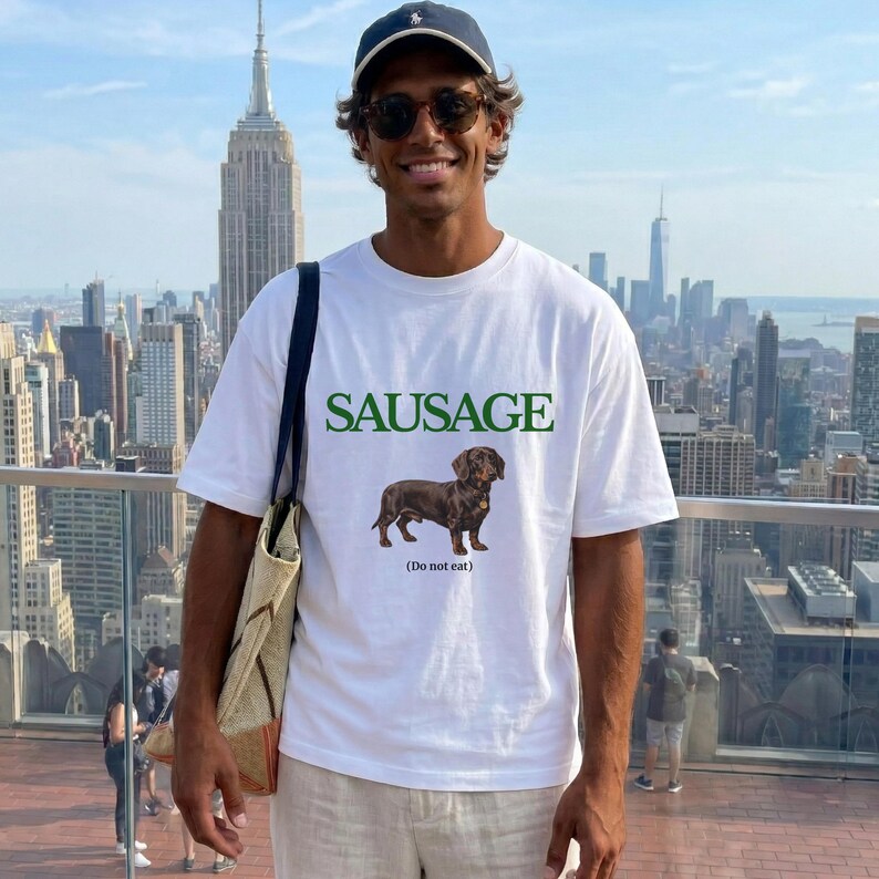 May include: White t-shirt with the word "SAUSAGE" in green block letters above a brown dachshund illustration. Below the dog is the text "(Do not eat)". The shirt is worn by a person in front of a city skyline.