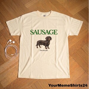 May include: Cream-colored t-shirt with the word "SAUSAGE" in green block letters above a brown dachshund illustration. Below the dog is the text "(Do not eat)". The shirt has a crew neck and short sleeves.