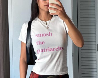 Pode incluir: T-shirt branco com a frase "smash the patriarchy" em letras rosa. A camisa tem mangas curtas e gola redonda. Uma bolsa preta de ombro é visível.